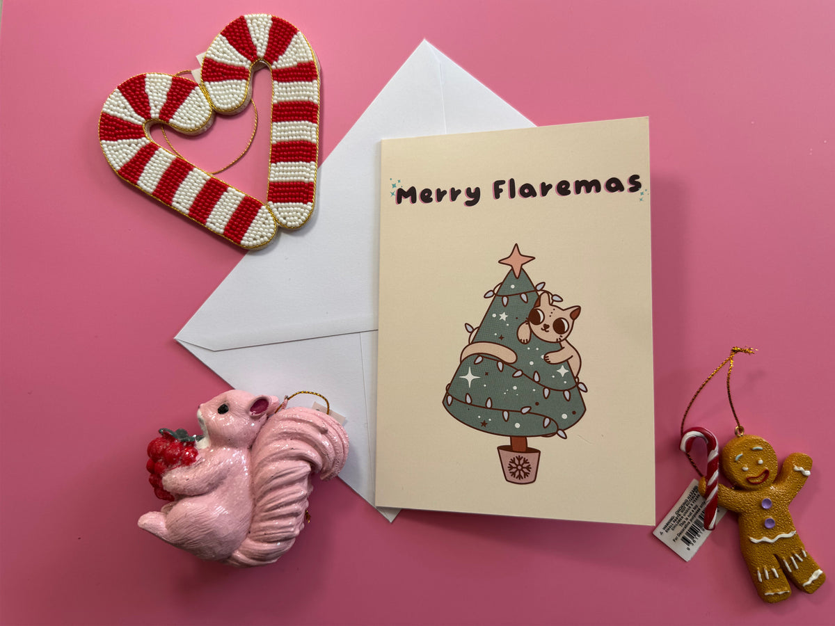 Merry Flaremas Christmas card with its envelope on a pink background surrounded by festive decorations.