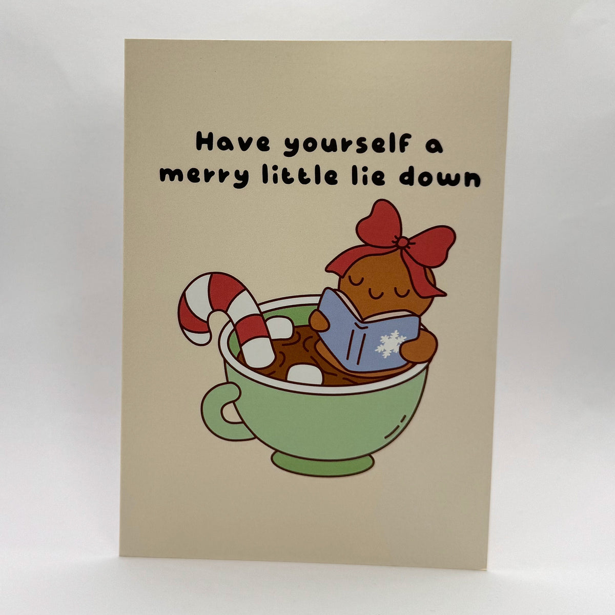 Have Yourself a Merry Little Lie Down A6 Christmas card with a cosy illustrated character holding a mug.