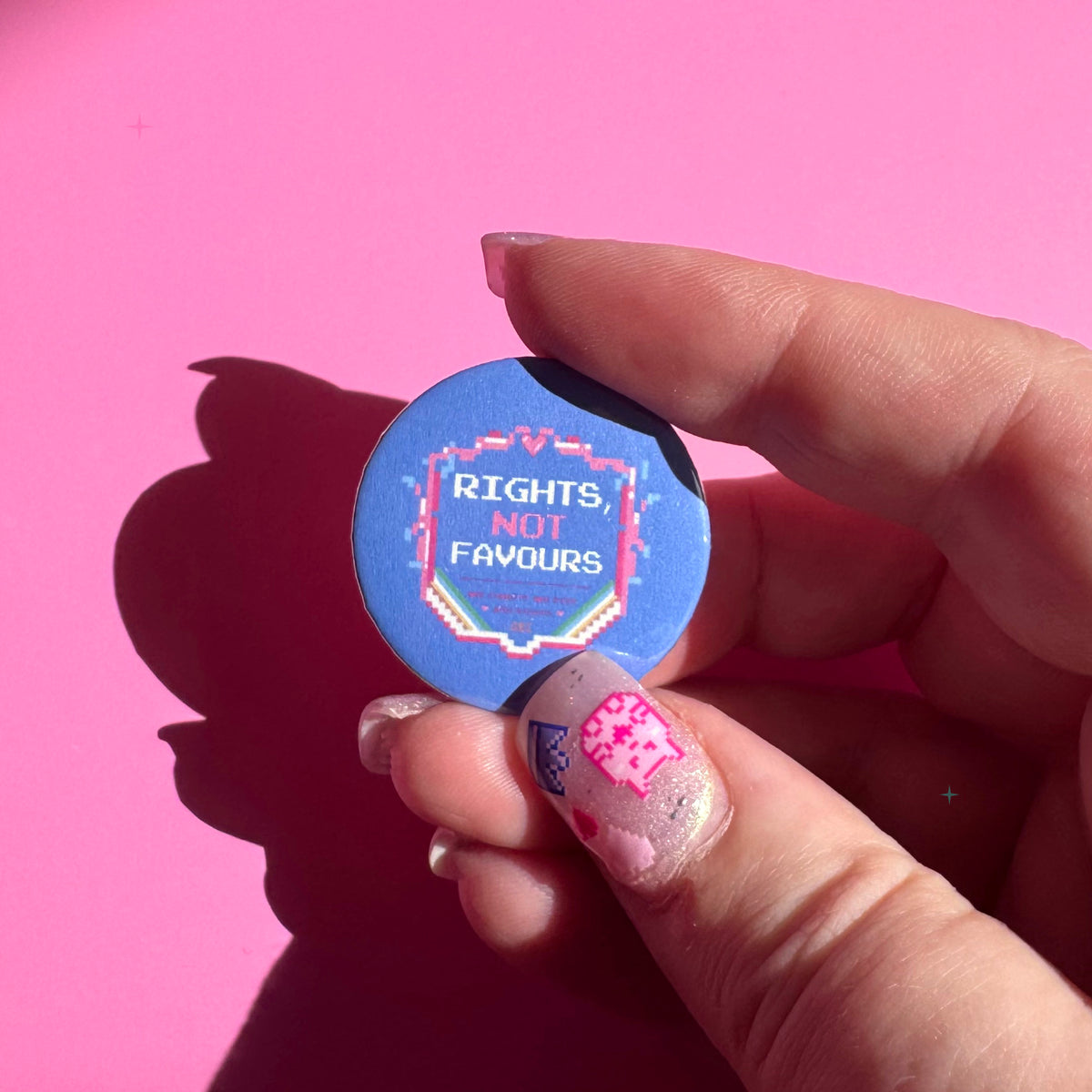 32mm blue Rights, Not Favours button badge held between fingers for scale